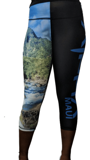 A person is wearing the Alpha Maui Iao Valley (3/4 Length) leggings, featuring a lush green landscape and flowing river print on the left leg. The right leg is adorned with blue vertical text reading "Maui." These leggings, reaching just below the knees, are perfect for beach apparel and embody the spirit of Aloha.