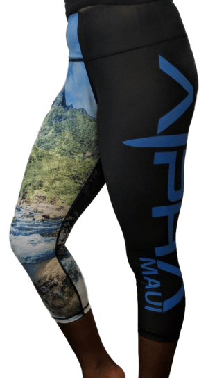 A person is wearing Alpha Maui Iao Valley (3/4 Length) black athletic leggings with "Aloha Maui" written in blue on the side. The leggings feature a scenic nature print on one leg, showcasing Hawaii’s greenery and a river under a blue sky. The person is standing in a partially angled side view.