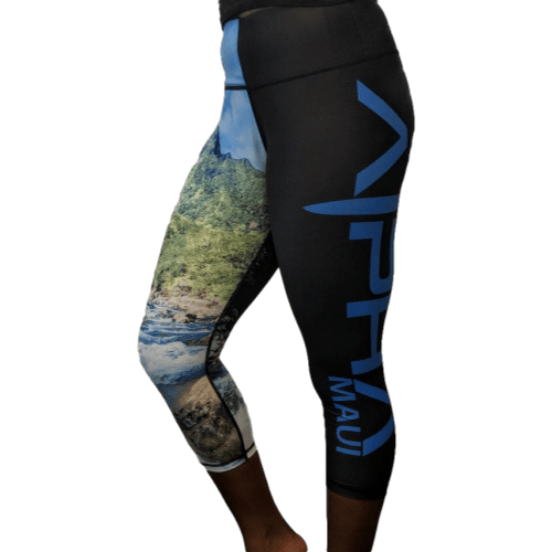 Someone is donning a pair of Alpha Maui Kepuhi Beach (3/4 Length) leggings in black, featuring distinctive blue text that reads "Maui" accompanied by a stylized logo. One leg highlights a scenic print depicting a lush green landscape with a river, capturing the essence of Aloha and the spirit of beachwear.