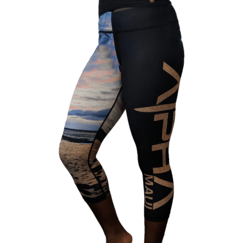 A person is wearing the Alpha Maui Kepuhi Beach 3/4 length leggings, which feature black and multicolored designs with the words "Alpha Maui." These beach apparel leggings exhibit a scenic print of a beach and sky on the left leg. Only the lower half of their body is visible against a transparent background.