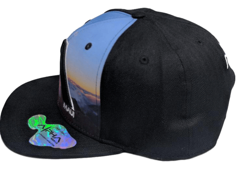 The Haleakala - Keiki Cap (Kid cap) features a black snapback design embellished with a vibrant rectangular patch on the front, showcasing a scenic sky and cloud pattern. A glossy holographic sticker boasting the word "Alpha" is affixed to the brim, adding a hint of Aloha spirit.
