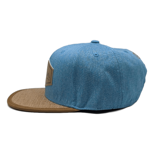 The Kam 2 cap, viewed from the left, showcases a unique design featuring a blue cap with a tan-brown bill and a patch on the front left side. Made of textured, fabric-like material and complete with eyelets for ventilation, its structured shape and casual style make it an ideal accessory for beachwear or an Aloha-inspired look.