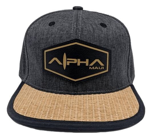 Introducing The La Perouse: a dark gray baseball cap with a tan woven brim. Adorning the front, you'll find a black hexagonal patch with gold embroidery that reads "Alpha Maui" in stylized letters, accompanied by "Maui" in smaller text. The front panel also features two small black ventilation holes, ideal for capturing those Aloha vibes from Hawaii.
