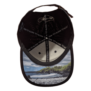 A black baseball cap called "The La Perouse" is pictured from the inside, showcasing stitching that reads "Aloha" and a printed signature. The underside of the brim features a scenic image of a Hawaii beach with trees, rocks, and water, making it essential beach apparel.