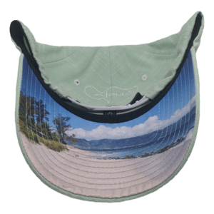A light green Sprecklesville baseball cap flipped to reveal the underside of the brim, showcasing a printed image of an Alpha Maui beach scene with white sand, trees, and a vibrant blue ocean beneath a clear sky. The inner top part of the cap features a small fish illustration and a tribute to the Ohana spirit.
