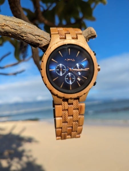 A wooden wristwatch with a black and blue dial featuring three small subdials is perched on a tree branch. The background showcases a sandy beach with the ocean and a clear blue sky, perfectly complementing your Alpha Maui beach apparel for that stylish Ohana vibe.