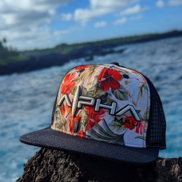 A hat with a tropical floral design and the words "Alpha Maui" sits on a rocky surface near the ocean. This stylish piece of beach apparel is set against a vibrant blue sky with scattered clouds and a blurred view of the coastline, capturing the true essence of Aloha.