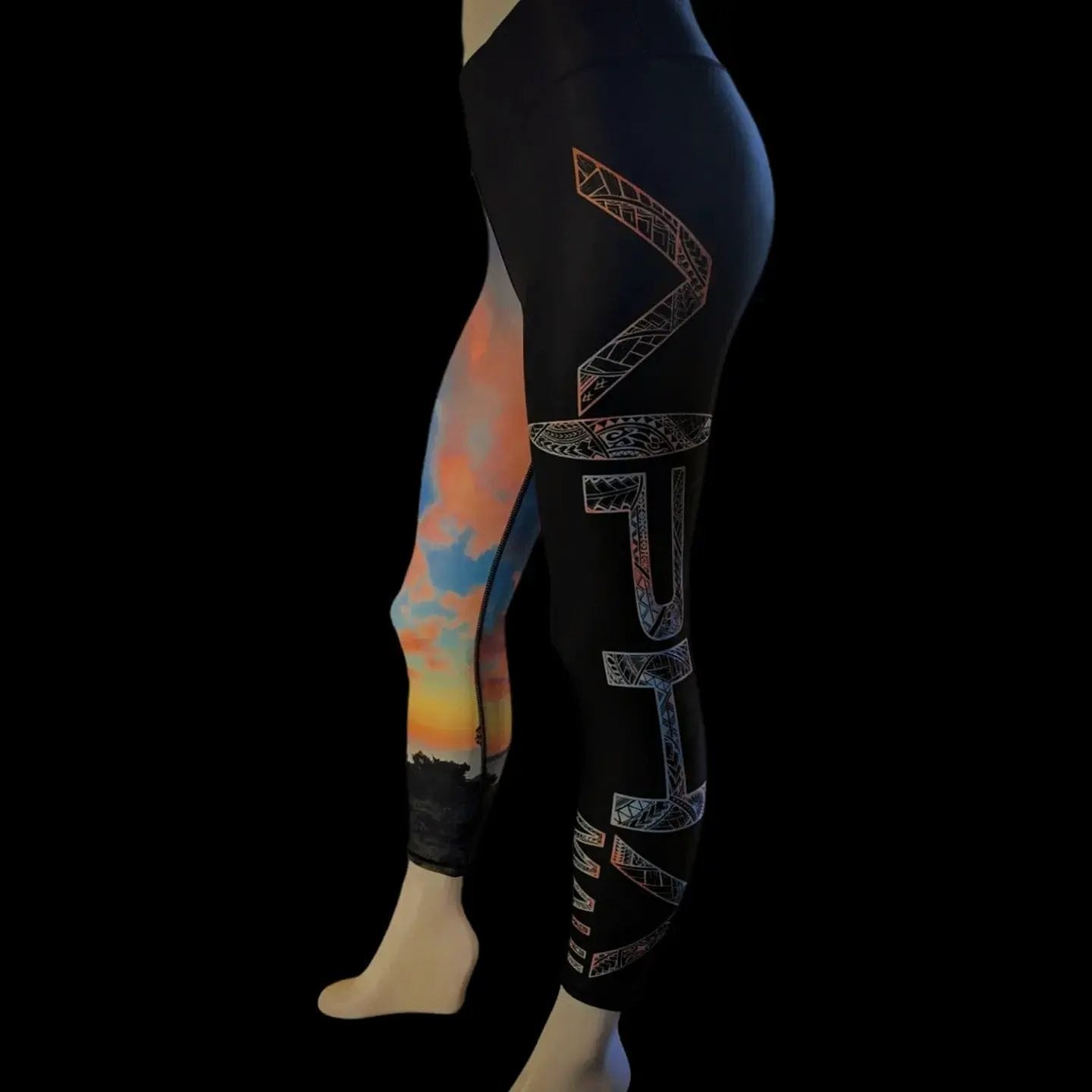 Side view of a person wearing leggings: The right leg is black with an intricate design featuring the word "native" vertically, while the left leg showcases a colorful sunset scene in shades of orange, blue, and pink. Perfect for beach apparel, the look evokes a sense of Ohana with a solid black background.