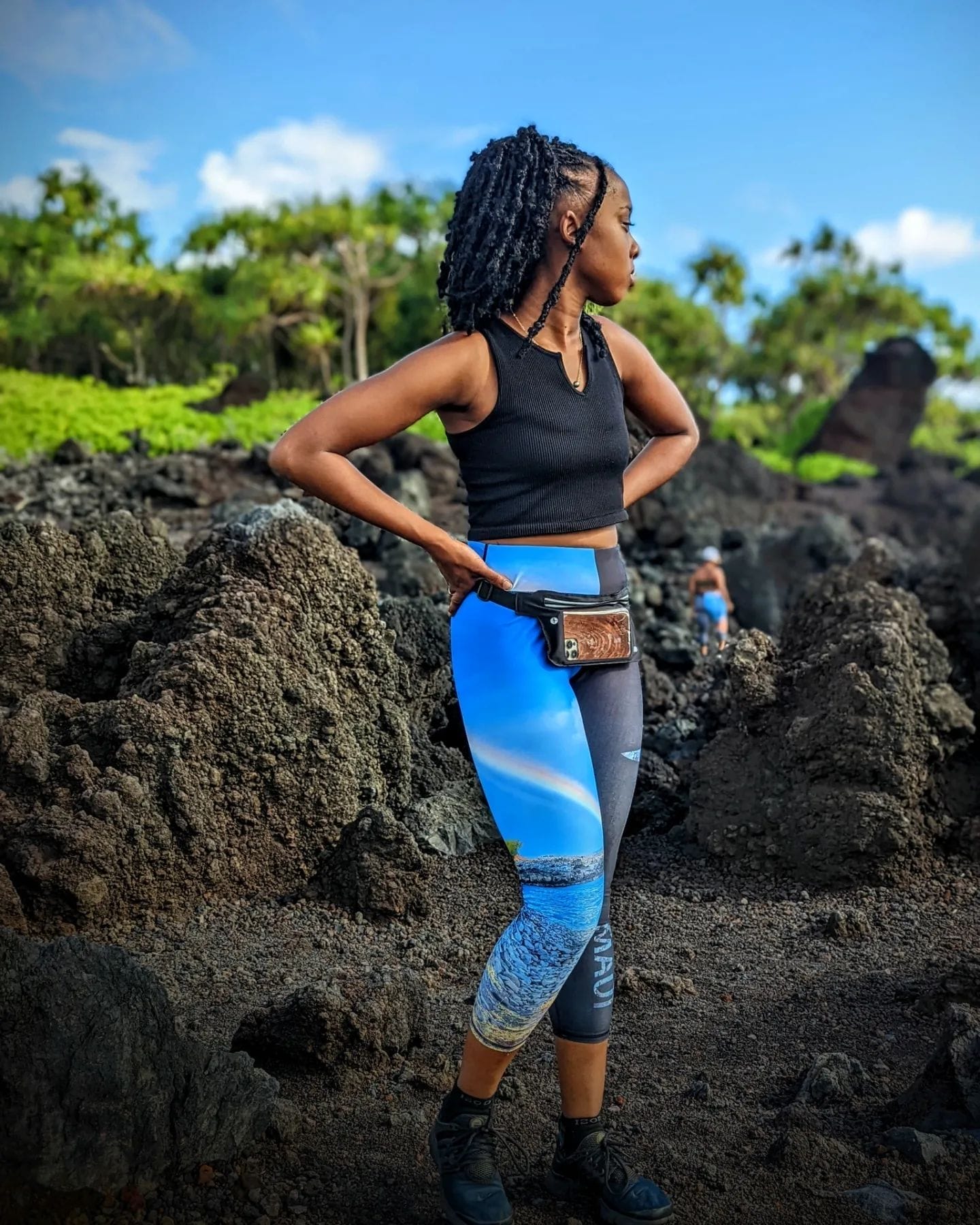 A woman in a black sleeveless top and blue patterned leggings, perfect for beach apparel, stands on rocky terrain with green foliage in the background. She has a small brown pouch around her waist and appears to be looking into the distance. The sky is clear and blue, radiating an Aloha vibe.