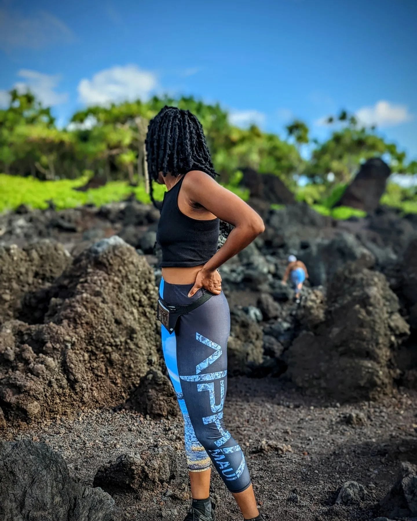 A person with dark, curly hair, wearing a black tank top and patterned leggings, stands on a rocky, volcanic terrain, posing with one hand on their waist. The lush, green landscape and a vivid blue sky are visible in the background, evoking an Aloha spirit.