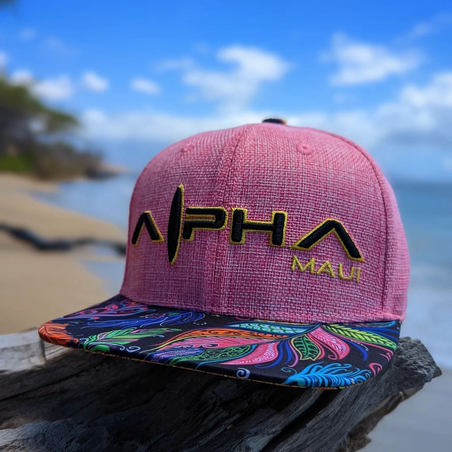 A pink Alpha Maui snapback cap with a vibrant, colorful brim featuring feather patterns sits on a piece of driftwood with a sandy beach and ocean waves blurred in the background under a bright blue sky, embodying the spirit of Aloha and Ohana.