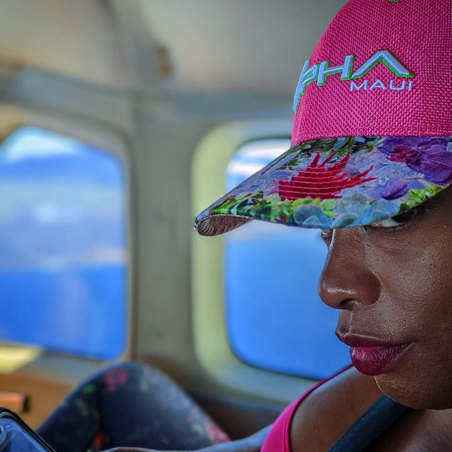 A person is seated by an airplane window, wearing a pink Alpha Maui cap with a floral brim and matching pink activewear. They are engrossed in using their smartphone, with a blue sky visible through the airplane window in the background.