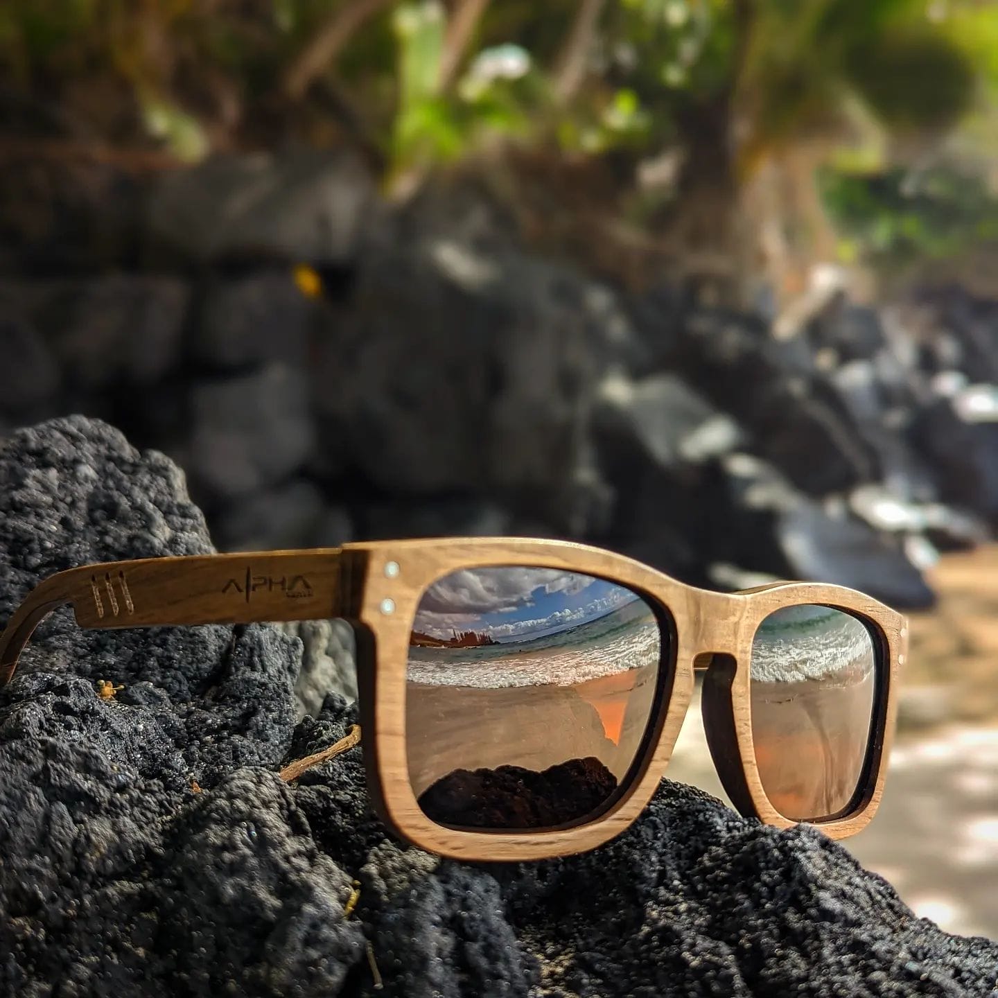 A pair of wooden sunglasses with reflective lenses rests on black rocks. The lenses capture a scenic Hawaii beach view with sand, rocky shores, and lush greenery in the background, under a bright, sunny sky—a perfect addition to your beach apparel.