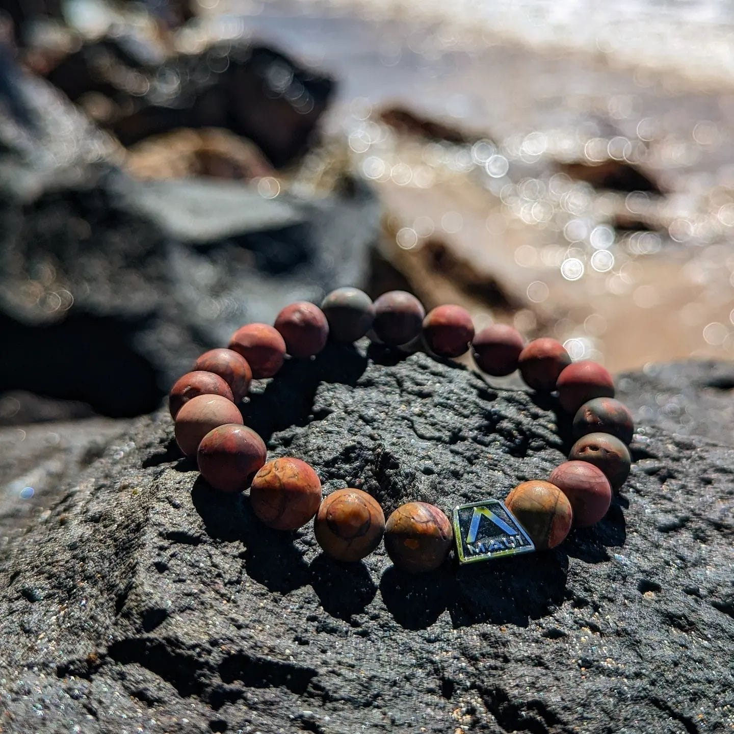 A beaded bracelet with earthy tones and a small triangular charm rests on a dark rock. In the background, the sunlight glimmers on the water and the rocky shore of Hawaii is visible but out of focus. This piece embodies the spirit of Ohana, reflecting Alpha Maui’s distinctive style.