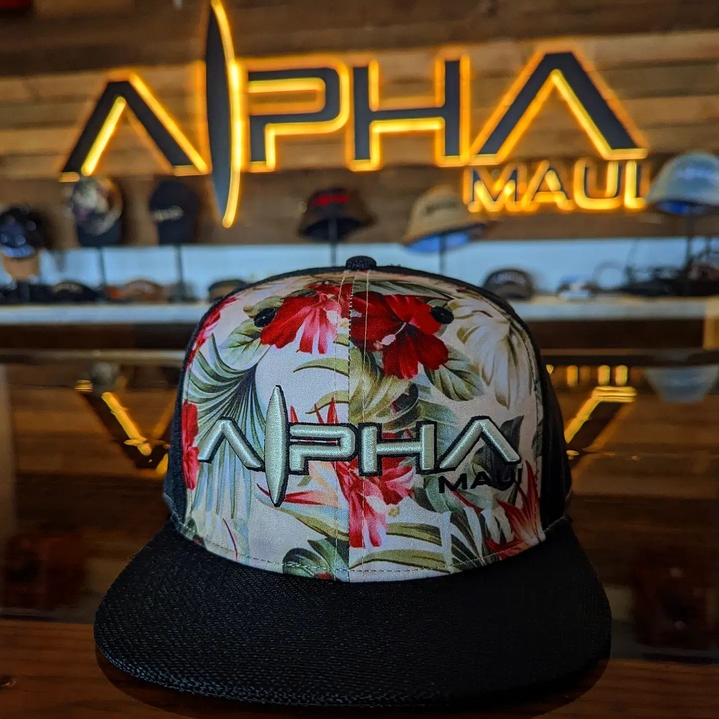 A floral print hat with "Alpha Maui" emblazoned on the front is displayed on a wooden surface. In the background, a wall features a lit "Alpha Maui" sign and various other beach apparel hats are visible.