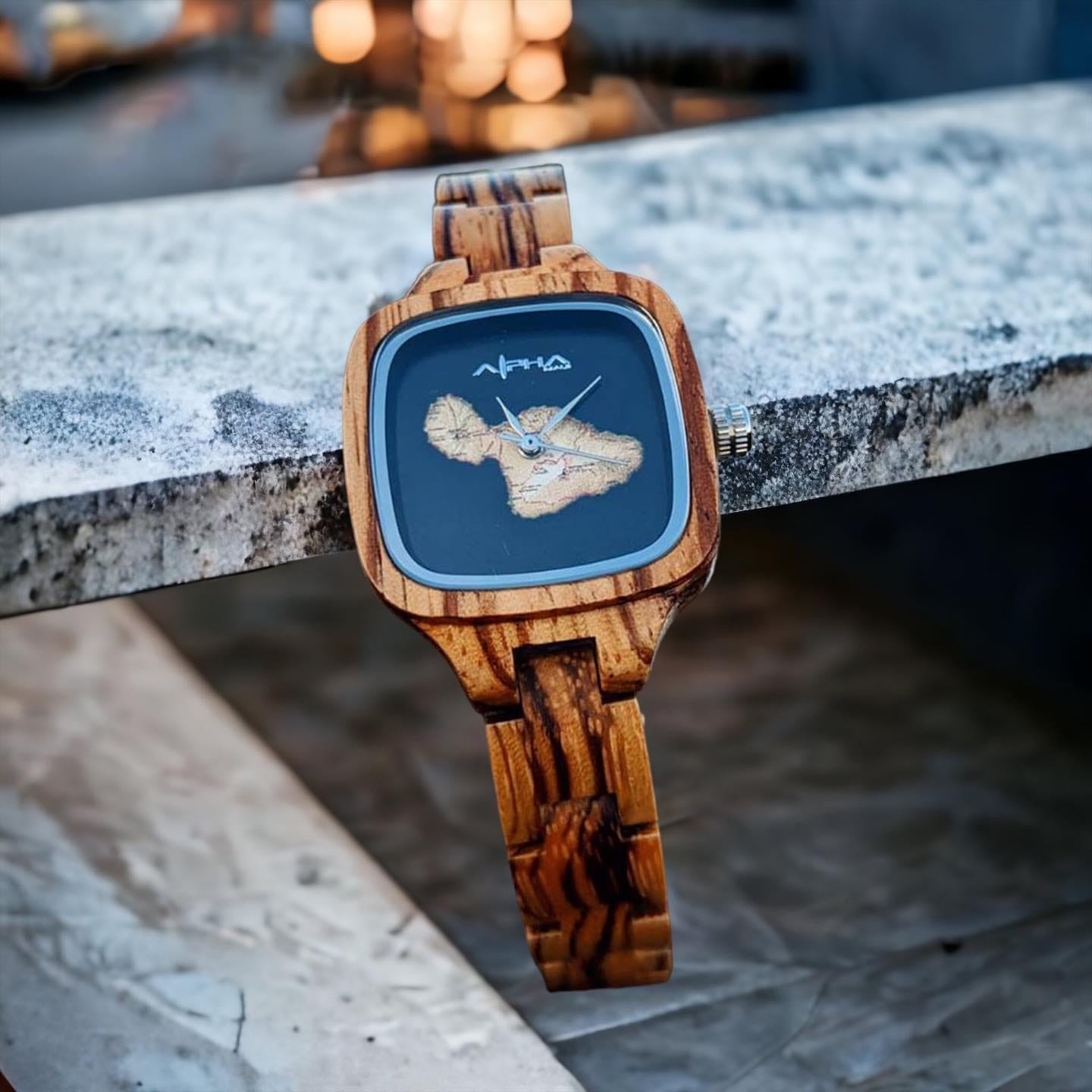 A wooden wristwatch featuring a unique design rests on a textured stone surface. The watch, ideal for complementing beach apparel, has a square-shaped face with a map detail of Hawaii on the dial and is made from intricately patterned wood, creating an elegant and earthy aesthetic reminiscent of Alpha Maui.