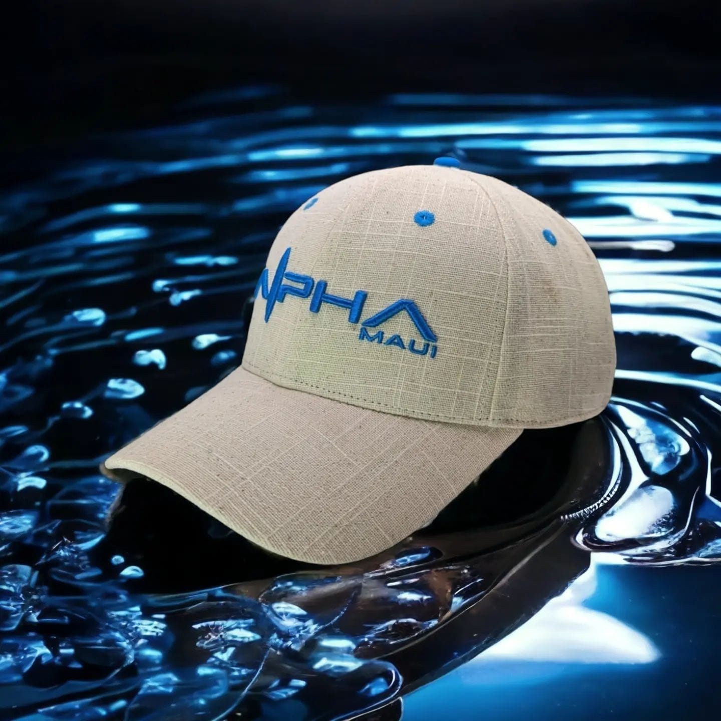 A beige baseball cap with blue embroidered text "Alpha Maui" and "Maui" is displayed on a reflective, watery surface. The cap features subtle textured patterns and blue ventilation holes. The dark background and water reflection enhance this beach apparel's design.