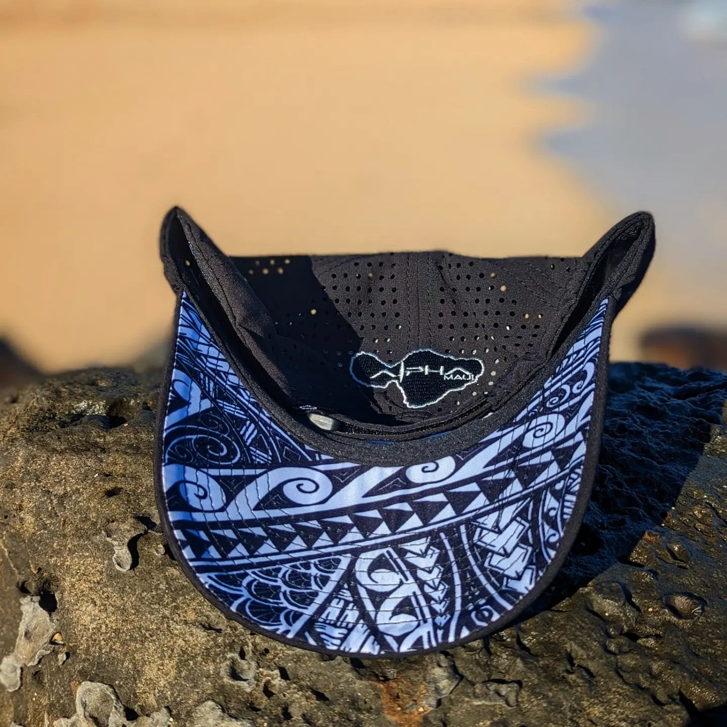 A black and blue visor with intricate white patterns on the brim lies on a textured rock. The interior includes a mesh material with a small embroidered logo. Perfect beach apparel, the backdrop features a blurred sandy beach and ocean reminiscent of Hawaii.