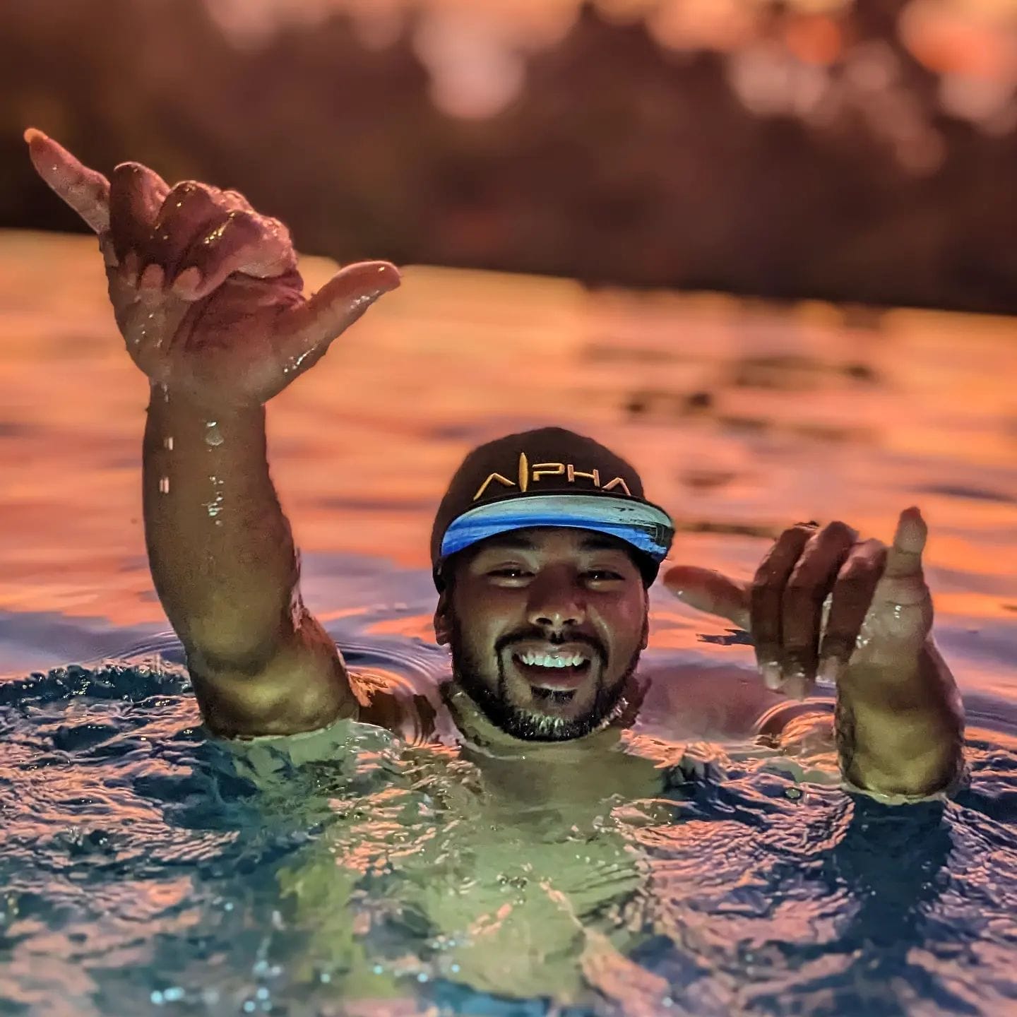         A man wearing a black baseball cap with the word "Alpha" enjoys swimming in a pool at sunset. He is smiling widely and making hand gestures, with both hands raised above the water. The background, slightly blurred, shows warm sunset colors and trees, giving it an "Aloha" vibe.