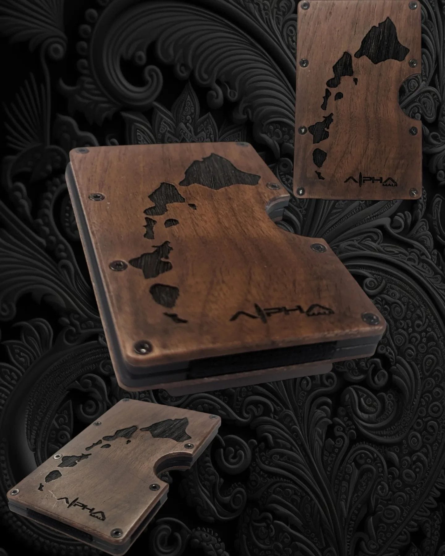 Dark wooden wallet-like accessory with etched designs depicting Hawaiian Islands. The accessory appears in three different views against a dark, intricate, paisley-patterned background. The word "Alpha Maui" is engraved on each piece, evoking the spirit of Aloha and complementing any beach apparel.