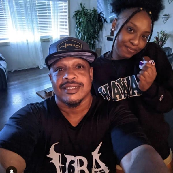 A man and a young woman take a selfie in a sunlit room with large windows. The man sports a black cap and shirt, while the young woman rocks a black "Hawaii" hoodie, her hair styled in a puff. Their warm smiles represent the spirit of Ohana.