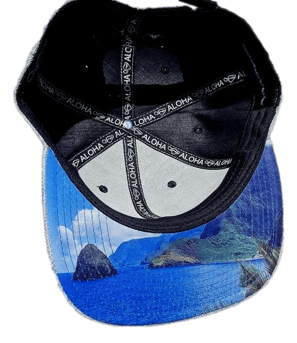 The Tinsman cap features a black and white design with the word "Aloha" repeated on its black interior straps. The underside of the brim displays a vibrant ocean scene complete with rocky cliffs and a clear blue sky, making it perfect beach apparel for anyone embracing the Alpha Maui spirit.