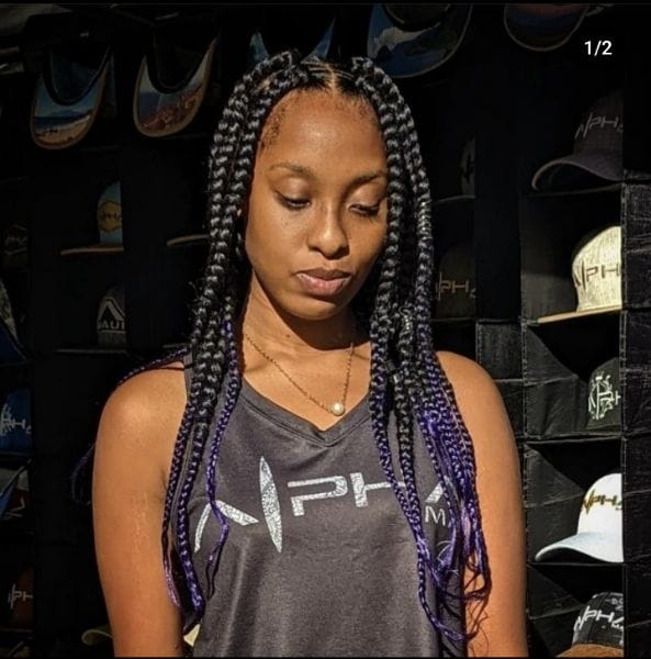 A woman with long braided hair, featuring purple extensions, is standing and looking down. She is wearing a sleeveless black top with the PHX logo and a delicate necklace. The background displays several baseball caps arranged on shelves, evoking a sense of Aloha.