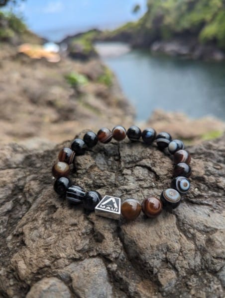 A beaded bracelet with dark and brown beads, centered on a rocky surface. The bracelet features a small tag with "Ohana Maui" inscribed on it. In the background, there is a blurred view of a water body, possibly a lake or river, with lush greenery reminiscent of Hawaii.