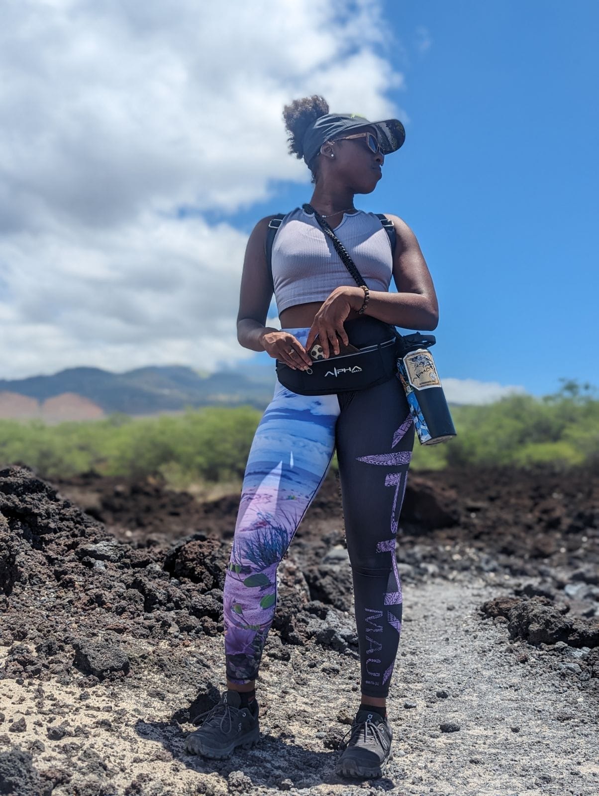 A person stands confidently outdoors under a clear blue sky in Hawaii. They're dressed in vibrant beach apparel, featuring colorful leggings and a sleeveless top. They wear sunglasses, a visor, and a waist pack with a water bottle attached. Rocky terrain and lush greenery are visible in the background.