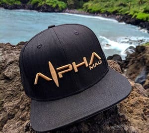 A black Baldwin Beach Bro cap with "ĀNHA" and "Maui" embroidered in gold rests on a rocky surface. The background features a scenic coastal view with lush green foliage, rugged rocks, and turquoise ocean water, epitomizing Alpha Maui beach apparel.