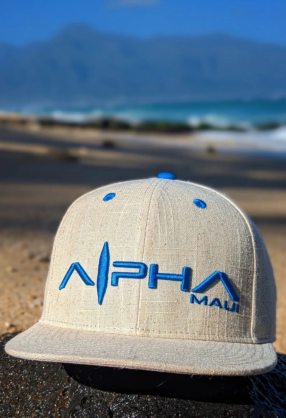 A beige baseball cap with a blue logo and the text "Alpha Maui" is perched on a sandy beach, blending seamlessly with Hawaii's blurred ocean waves and distant mountains in the background.