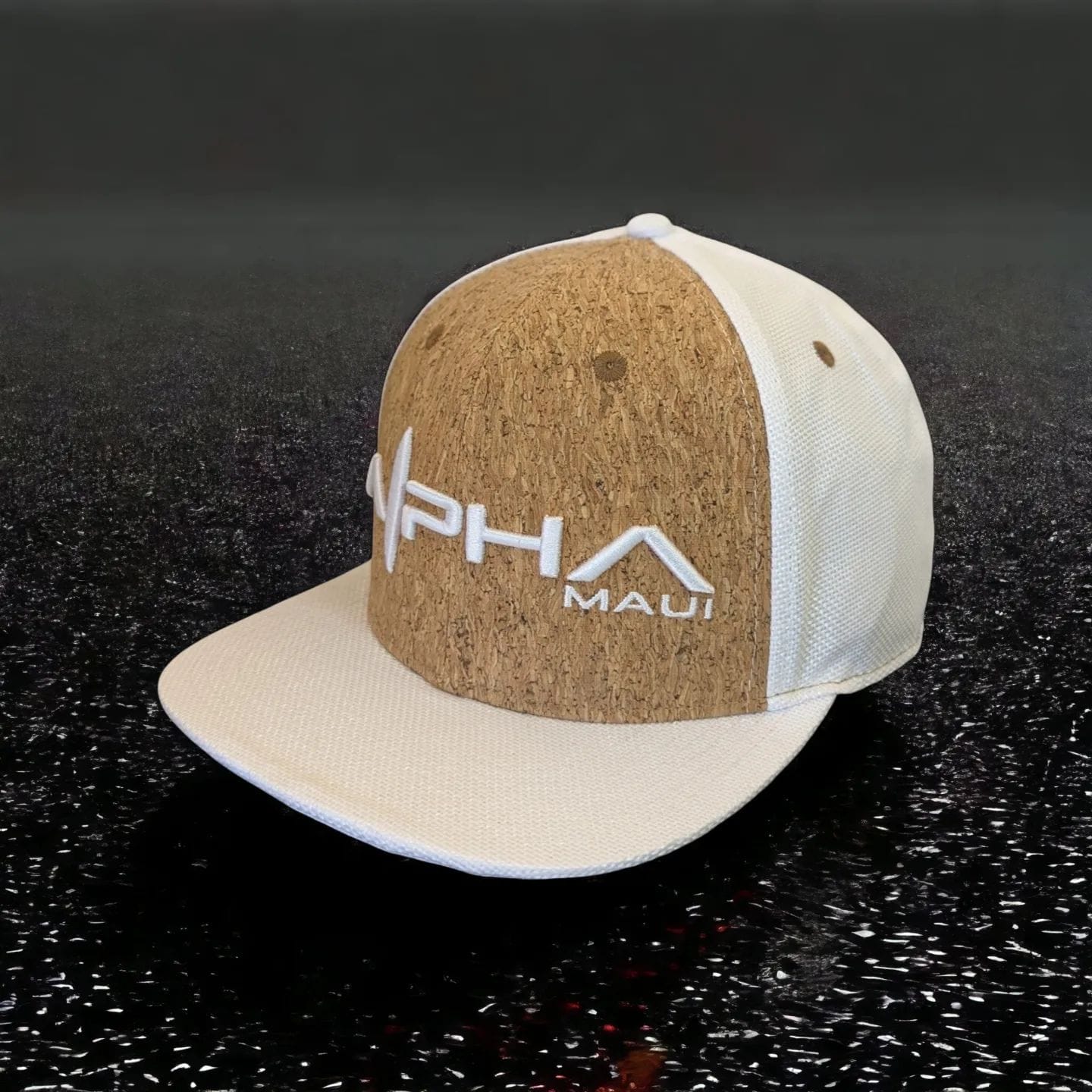 A two-tone baseball cap featuring a cork front panel with the text "Alpha Maui" and a stylized roof design embroidered on it. Perfect for beach apparel, the rest of the cap is white, with a flat brim and white mesh on the sides and back, set against a dark textured background.