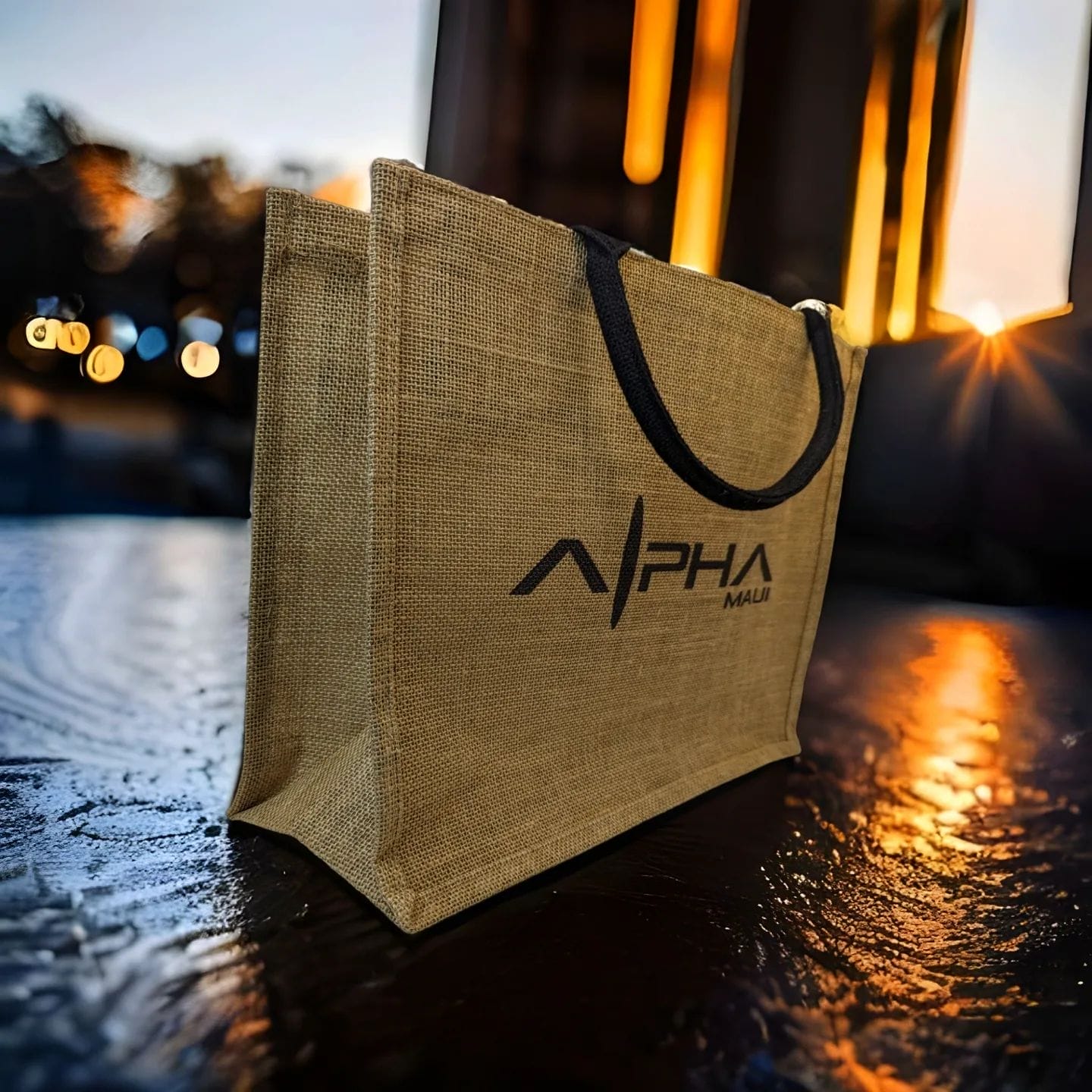 A jute tote bag with "Alpha Maui" printed on it in black text sits on a reflective surface. The background features blurry, illuminated buildings and trees, with the sun setting, casting a warm glow and deep shadows that evoke a sense of Aloha spirit from Hawaii.