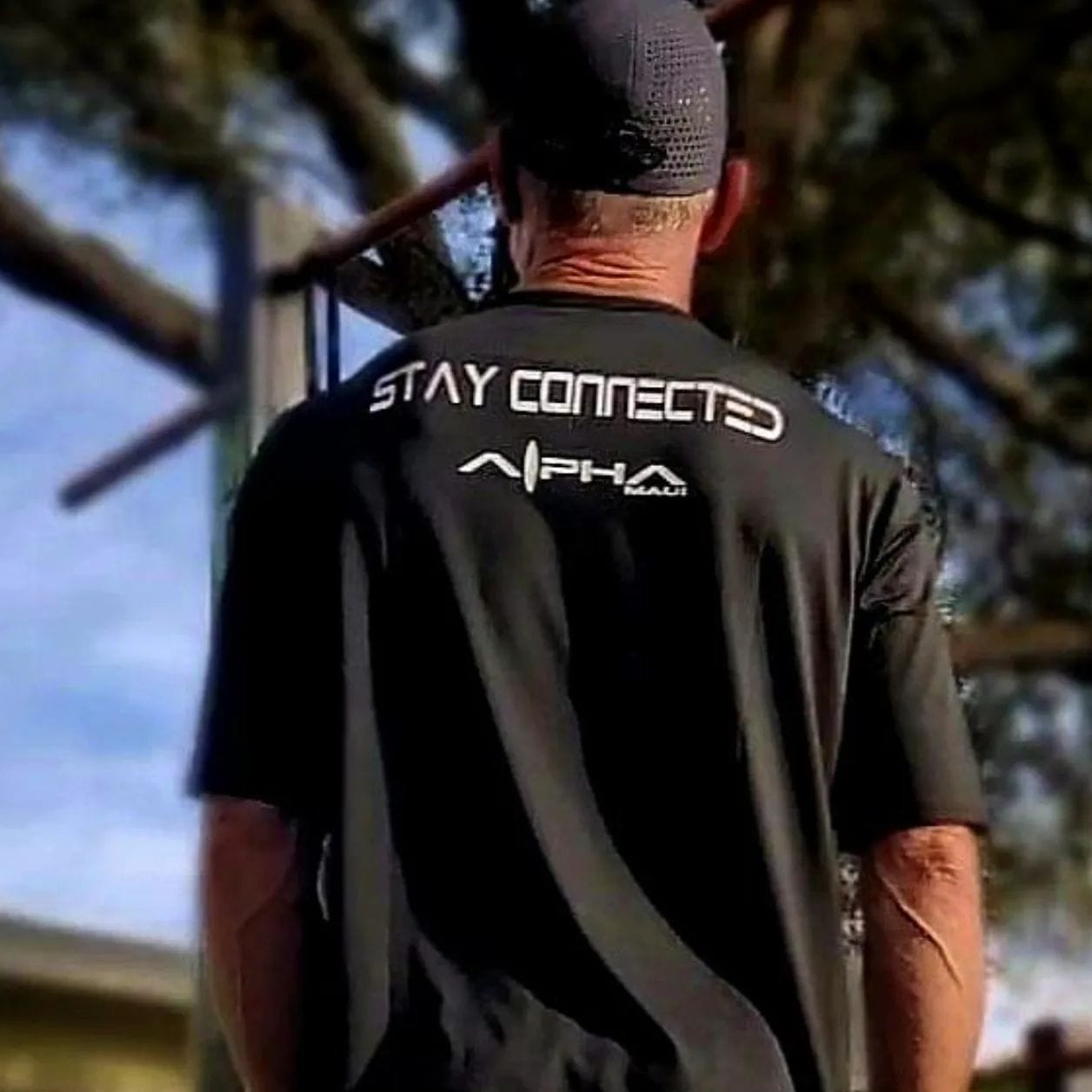 A person wearing a black shirt with the phrase "STAY CONNECTED" and "Alpha" branded below it stands facing away. Trees and a clear blue sky create a picturesque backdrop, giving off Hawaii vibes reminiscent of beach apparel and the spirit of Aloha.