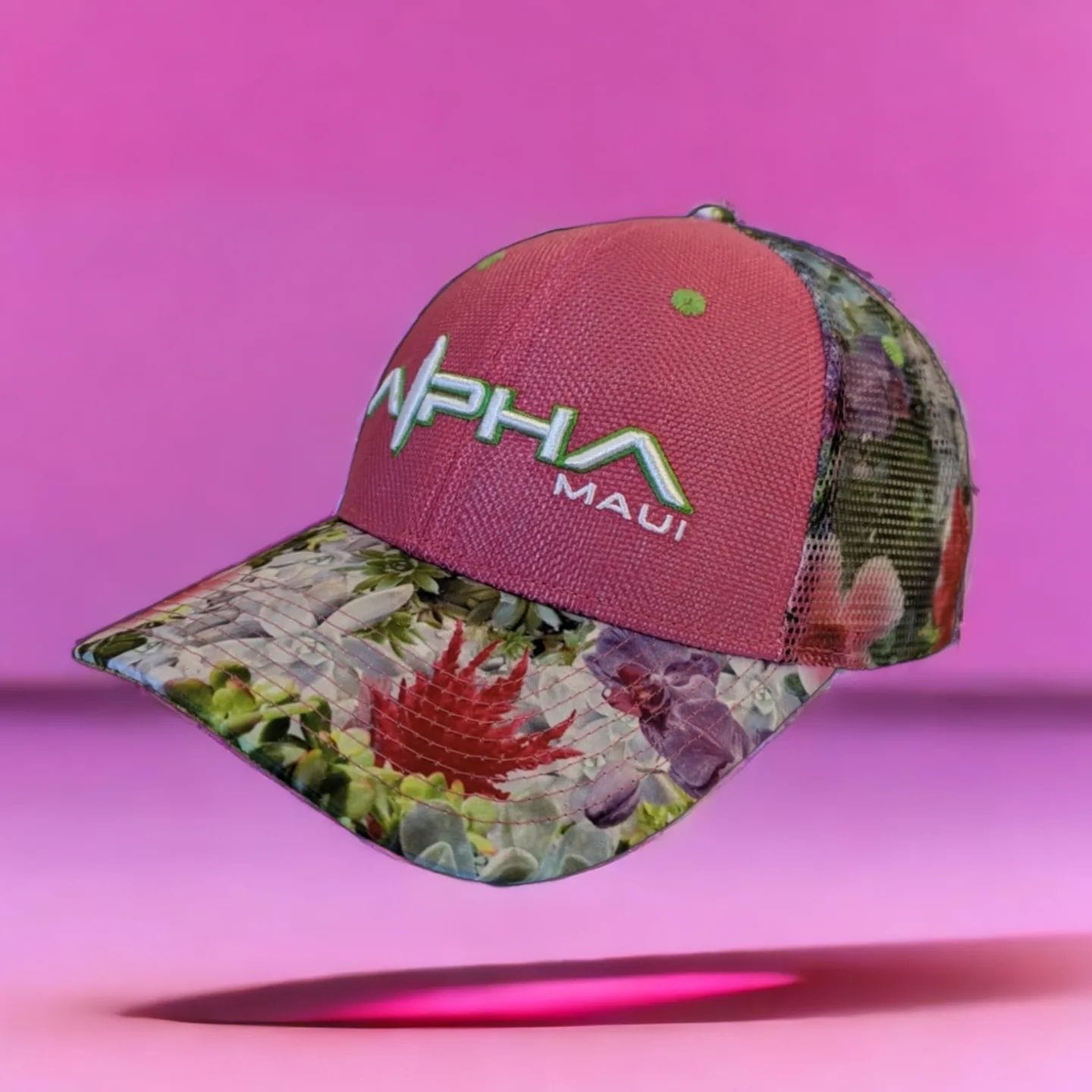 A colorful baseball cap featuring a combination of floral and tropical patterns on the brim and back, with a solid pink front panel. The front has "Alpha" and "Maui" embroidered in gradient blue and green thread, bringing a touch of Aloha vibes. Perfect for your beach apparel collection.