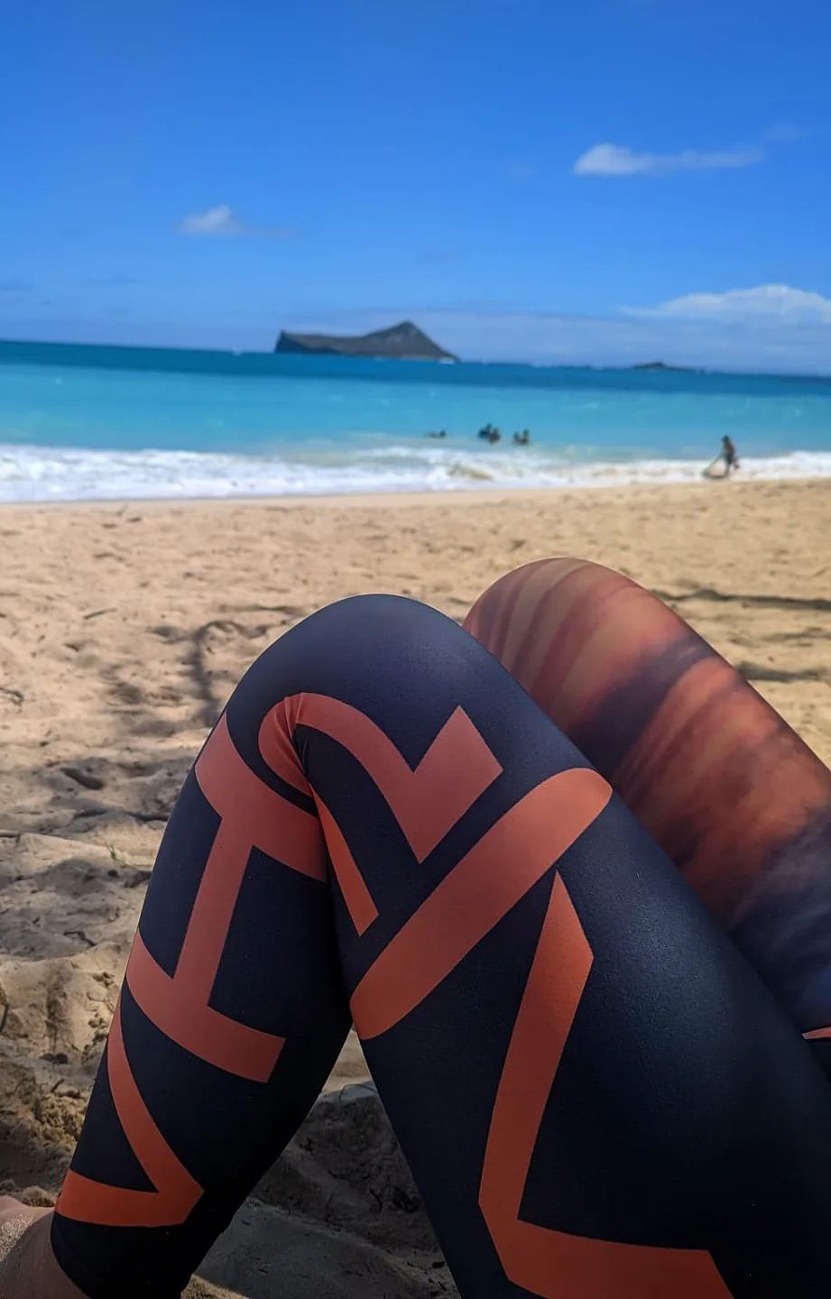 Close-up of a person sitting on a sandy beach in Hawaii, displaying their legs in stylish beach apparel—black leggings with orange geometric patterns. The background shows a turquoise ocean, a small island, and a clear blue sky with scattered clouds. Other beachgoers are visible in the distance enjoying the Aloha vibe.