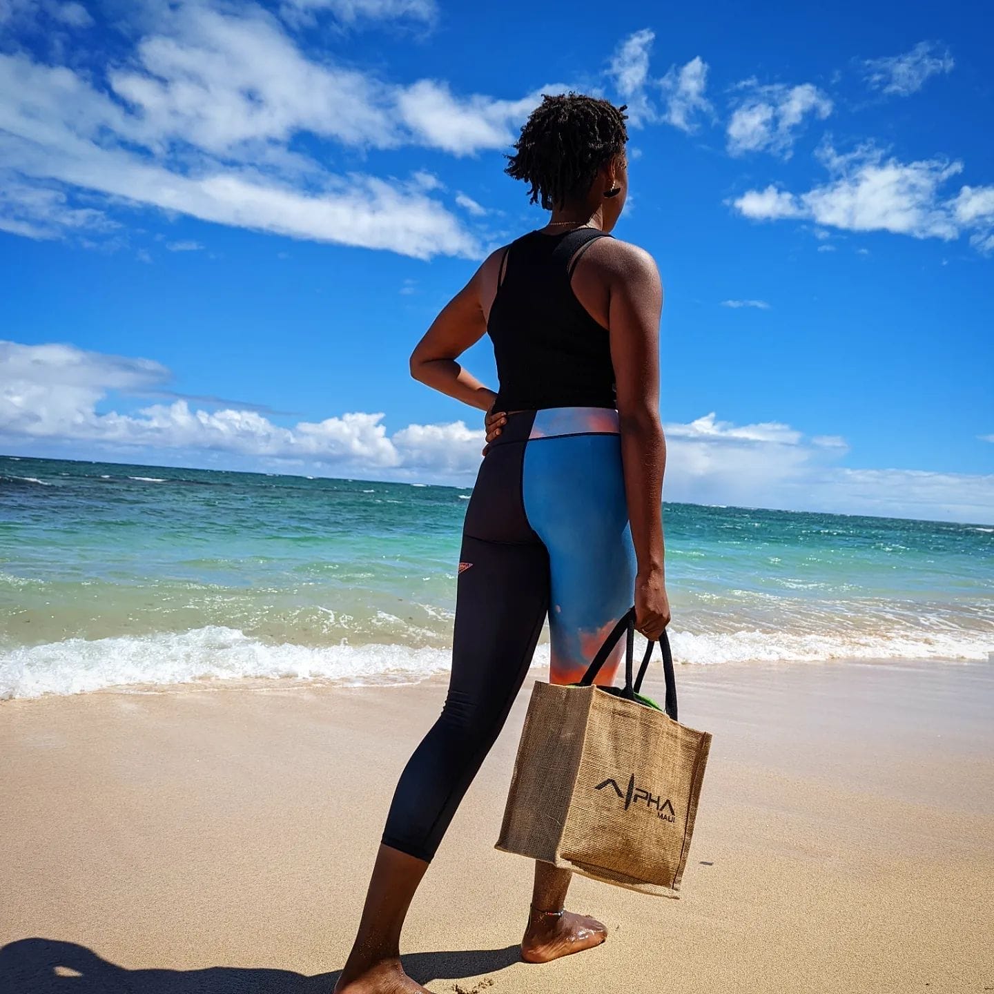 A person stands on a sandy beach in Hawaii, facing the ocean. They are wearing a black tank top and blue and orange leggings, holding a tan tote bag with "PHA" written on it. The sky is blue with a few scattered clouds, capturing the perfect Aloha spirit.