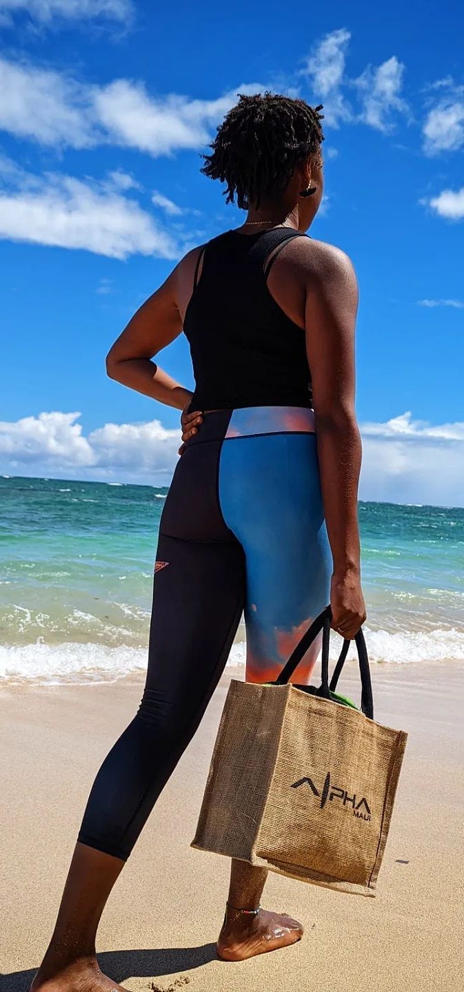 A person stands on a sunny beach facing the ocean, holding an Alpha Maui burlap tote bag. They are wearing a black sleeveless top and colorful leggings with shades of blue and pink. The calm sea and bright blue sky with a few clouds create the perfect Aloha vibe in the background.