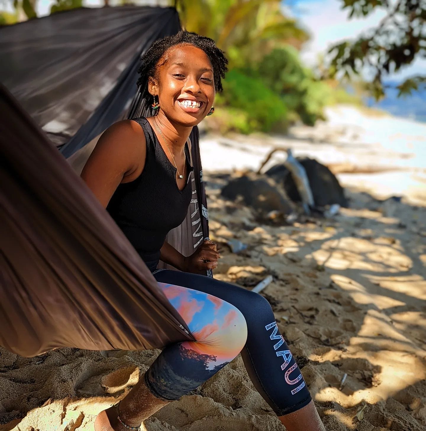 A woman with short hair sits smiling on a beach in a dark sleeveless top and leggings that say "Maui." Her left hand holds a strap of a hammock, and there is greenery and the ocean in the background. Sunlight casts a warm glow on her, capturing the Aloha spirit of Hawaii.