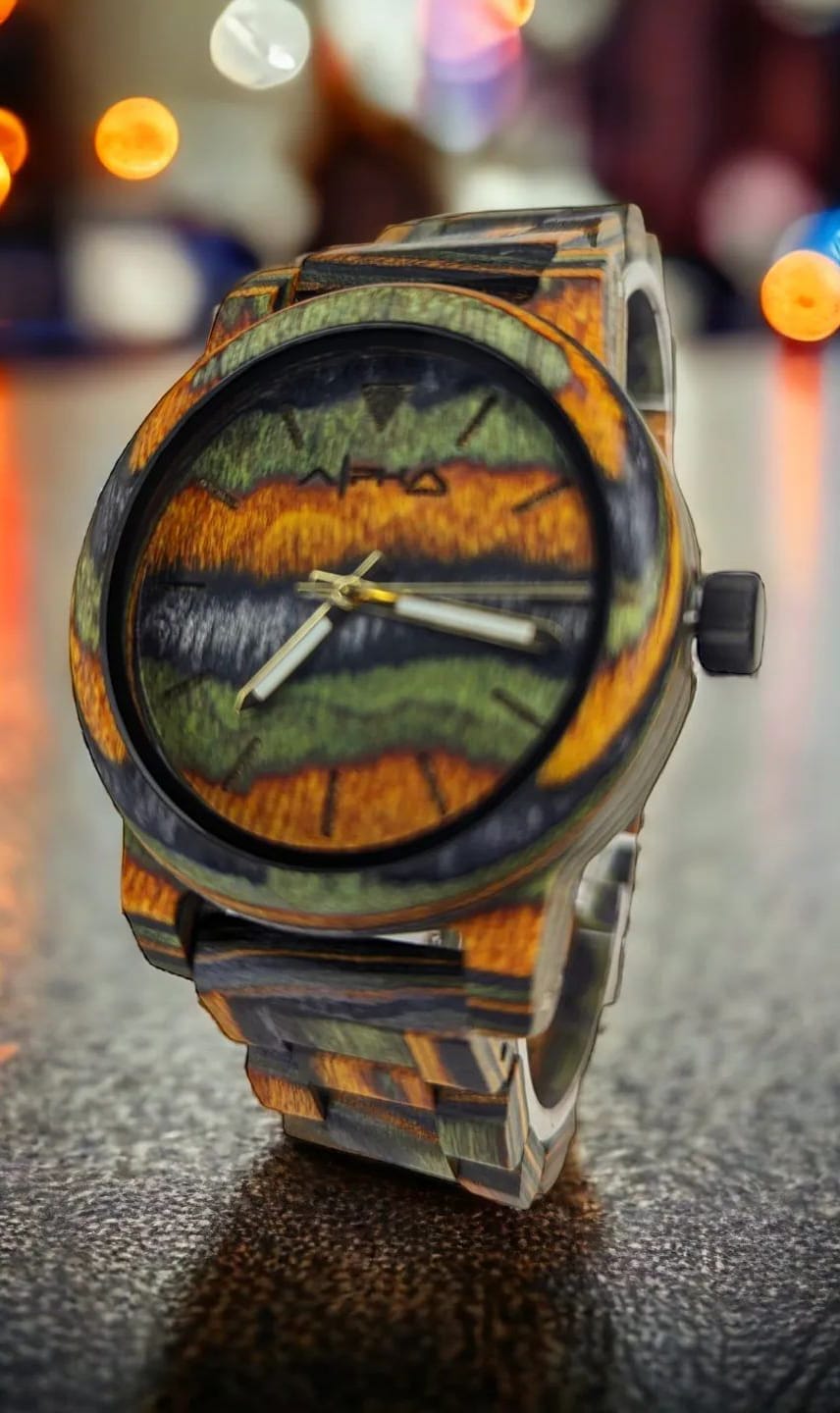 A close-up of the Alpha Maui wristwatch featuring a vibrant, multicolored matte finish with a camouflage-like pattern. The watch has a round face with golden and black tones, simple hour and minute hands, and a matching multicolored strap. The background is blurred with bokeh lights, reminiscent of beach apparel in Hawaii.