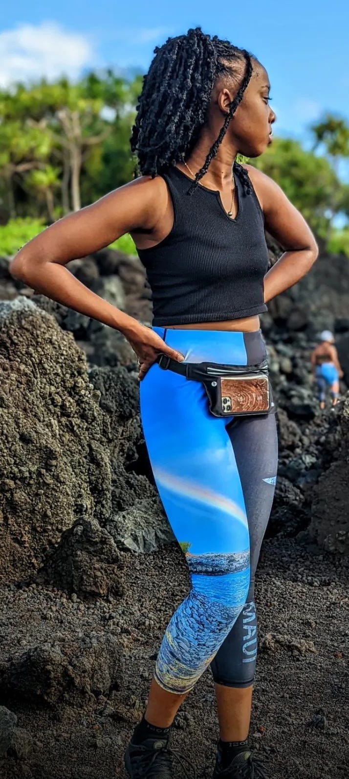 A person wearing a black sleeveless top and vibrant leggings, featuring a beach scene, stands on rocky terrain. They have braided hair, and a phone is attached to the waistband of their leggings. Lush greenery is visible in the background, evoking an Aloha spirit reminiscent of Hawaii's natural beauty.