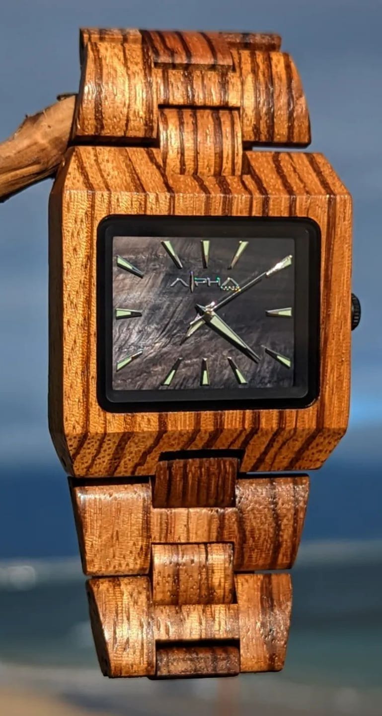 A wooden wristwatch with a square face, displaying a black dial and green glow-in-the-dark hands and hour markers. The case and band are made from striped natural wood, creating a rustic and organic appearance reminiscent of Hawaii. The background is a blurred outdoor scene.