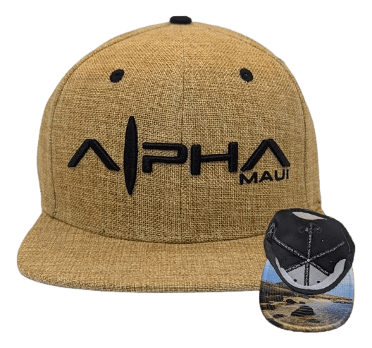 The Hemped Out baseball cap features a tan color with black embroidered text reading "Alpha Maui" on the front. The underside of the brim showcases an illustrated coastal landscape with rocks, ocean, and sky, perfectly capturing the spirit of Hawaii.