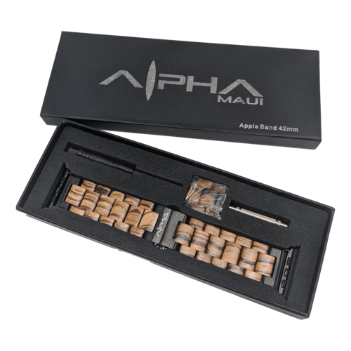 An Alpha Maui Band - Zebrawood 42mm set is displayed in its opened packaging. The box includes a wooden link watch band designed for a 42mm Apple Watch, extra links for size adjustments, a small screwdriver, and a pin removal tool. The lid is labeled “Alpha Maui” and "Apple Band 42-49mm," invoking the spirit of Aloha from Hawaii.