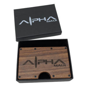 An Alpha Clip wooden wallet, engraved with the brand name "Alpha Maui," rests inside an open black box that also features the "Alpha Maui" branding on its lid. The wallet boasts a minimalist design and is partially recessed into the box, encapsulating the essence of Hawaii and Aloha spirit.