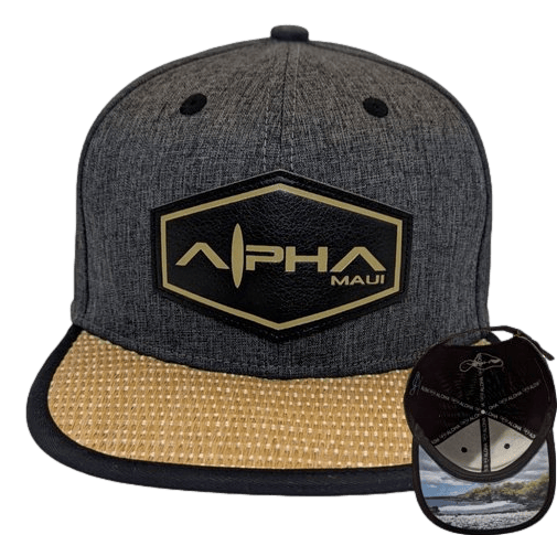 The La Perouse is a grey baseball cap featuring a hexagonal black and gold logo that reads "Alpha Maui" on the front. The brim has a woven texture in tan color. A circular tag attached to this beach apparel depicts a scenic beach scene with trees and water, all set against a black background, adding a touch of Aloha.