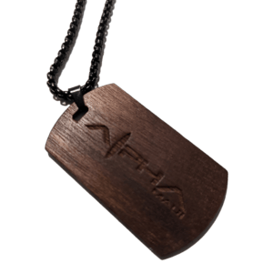 The Alpha Tag is a wooden dog tag pendant featuring a carved design of Alpha Maui's minimalist mountain logo. It hangs from a black chain and has a distinctive rectangular shape with a subtle curve at the bottom and an angular notch at the top, making it the ideal accessory for your beach attire.