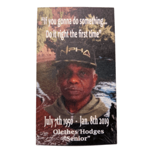 A memorial card showcasing a portrait of a man wearing a black cap and green jacket. The serene background features water and lush greenery. Above, the text reads, "If you're gonna do something, do it right the first time." Below are his name and dates: July 7th, 1950 - Jan 8th, 2019. Ohana forever. Product Name: Alpha Tag.