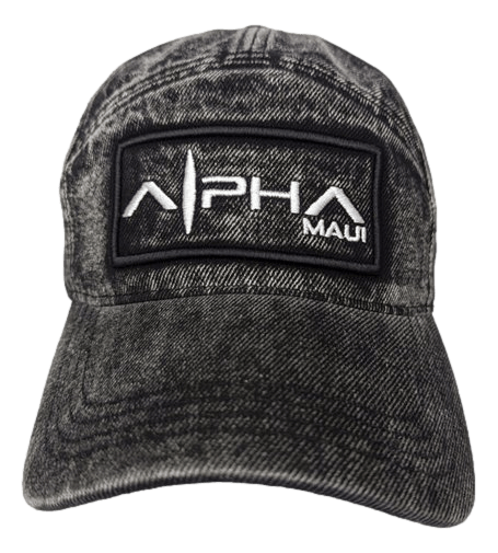 The Alpha Maui Zuppke is a dark, slightly worn denim cap featuring a black rectangular patch with the words "Alpha Maui" in white and a graphic design resembling a wave or mountain peak. The word "Aloha" is also embroidered in white on the patch.
