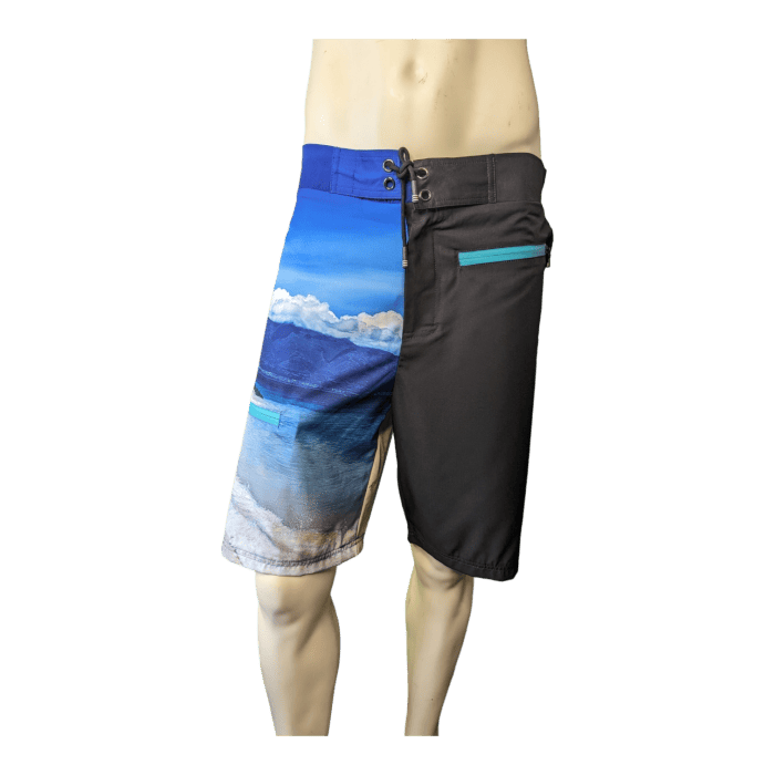 A pair of knee-length "Lava Flow" board shorts on a mannequin, showcasing a scenic beach and ocean print on the left leg and solid black fabric on the right leg. This Hawaii-inspired design features a blue and turquoise ocean scene with mountains in the background, and includes a teal zippered pocket on the right side. Perfect for any beach apparel collection.